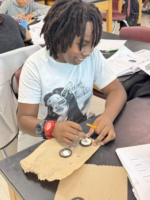 Student in classroom, smiling, working on craft project with small round items and paper.