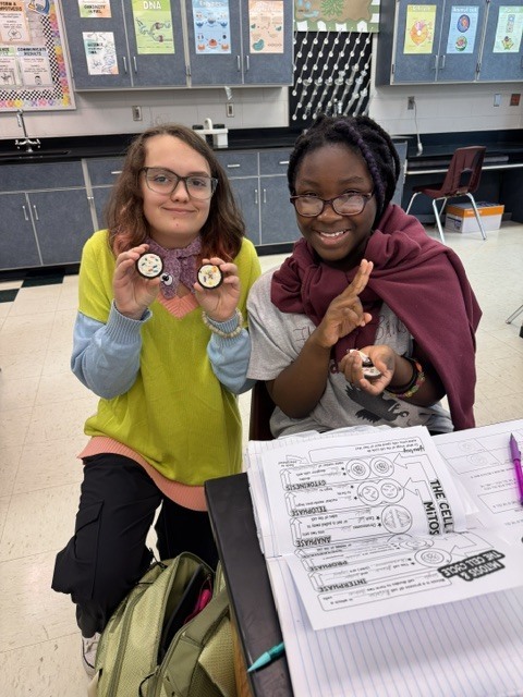 Two students sitting at a table with paper and pens. One holds small items in hand.