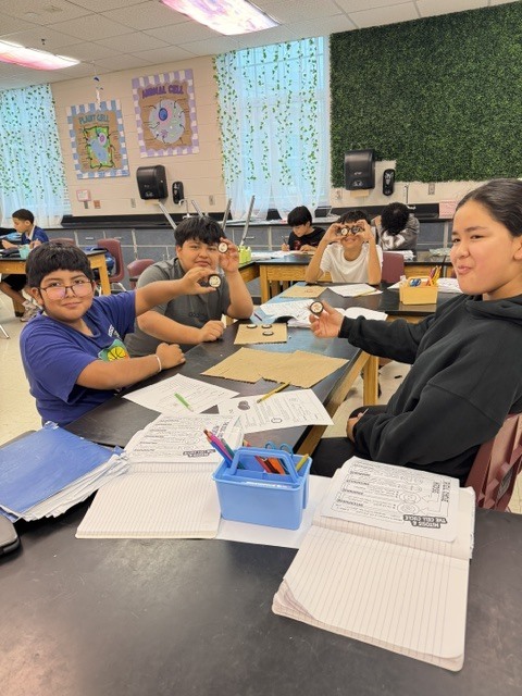 Four students at a table, seated with paper, pencils, and a small object, in a classroom with white blinds and green wall.