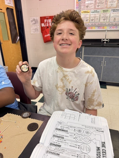 A boy sits at a table in a classroom, holding a small object and smiling.