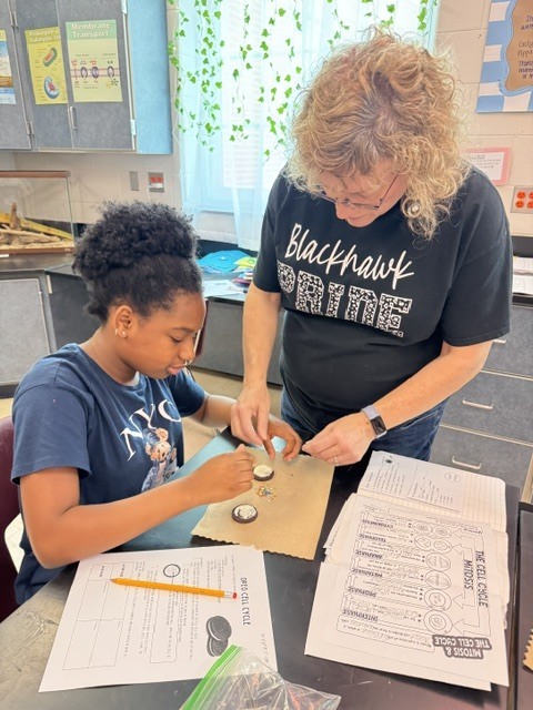 Two individuals seated at a table, working on a project. A student with curly hair wearing a blue shirt, a woman with glasses wearing a black shirt.