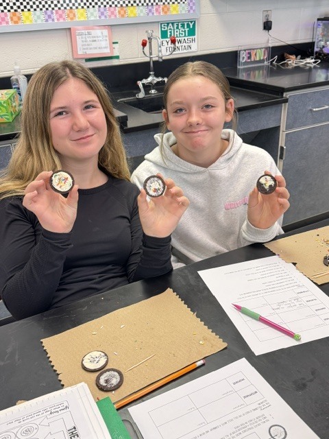 Two girls in a classroom hold small objects. One holds a circle, the other a square. Papers, pencils, and a pen on the desk.