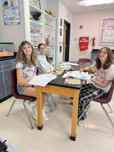 Three students seated at a table with papers, pens, and a fire extinguisher in the background.