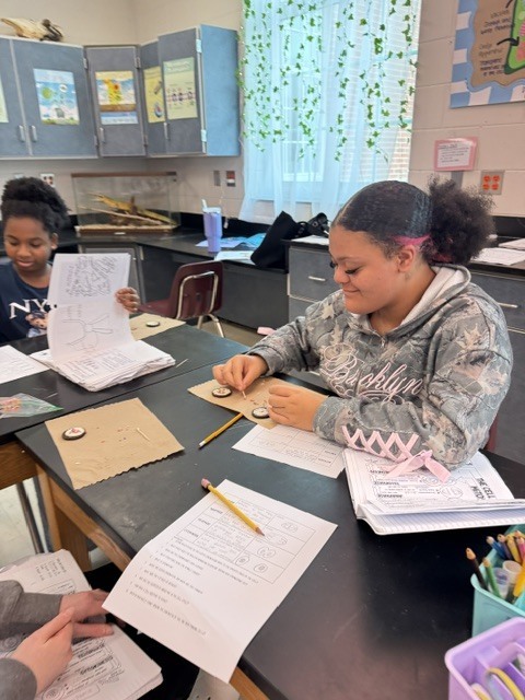 Two students working at desks with papers, pencils, and other materials in a classroom.