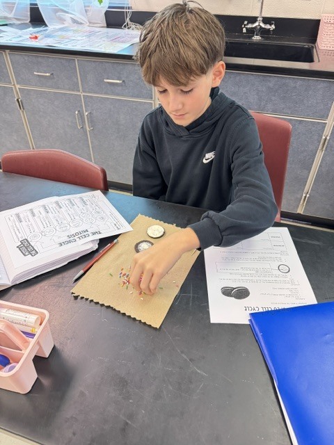 A student sits at a table, working with paper, coins, and a small pouch.