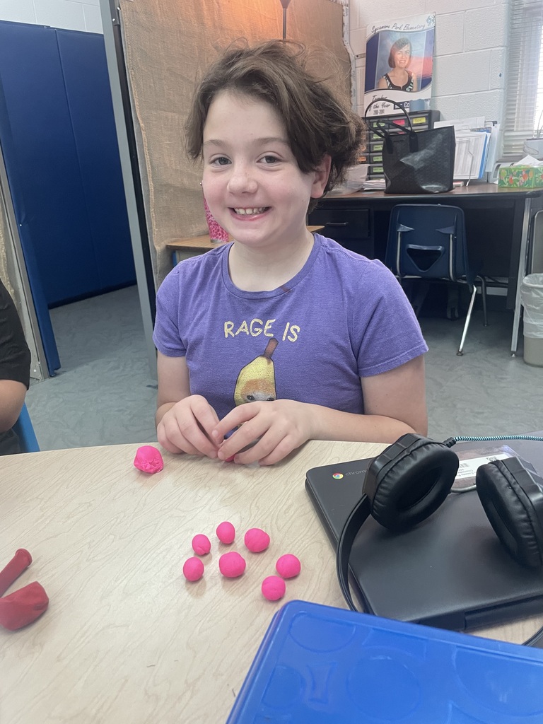 A girl in a purple shirt sits at a desk with pink objects and headphones.