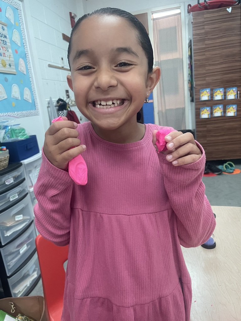A smiling child in pink holds up two pink objects in both hands. Behind her is a room with shelves and storage drawers.