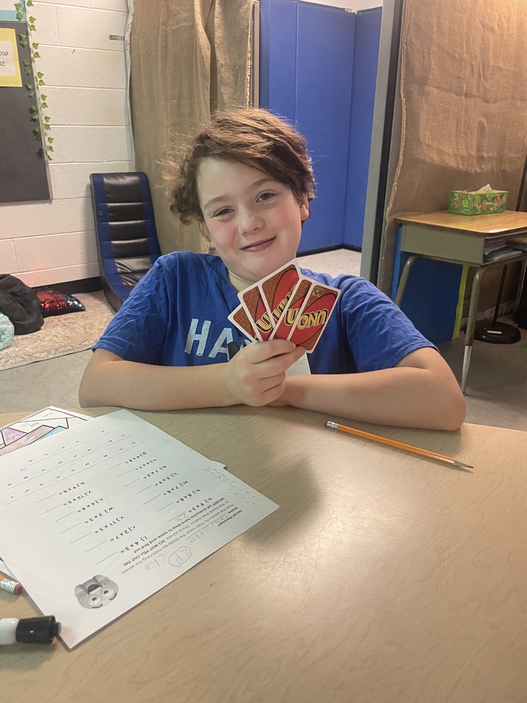 Boy holding cards, sitting at a desk with a paper, pencil, and marker.