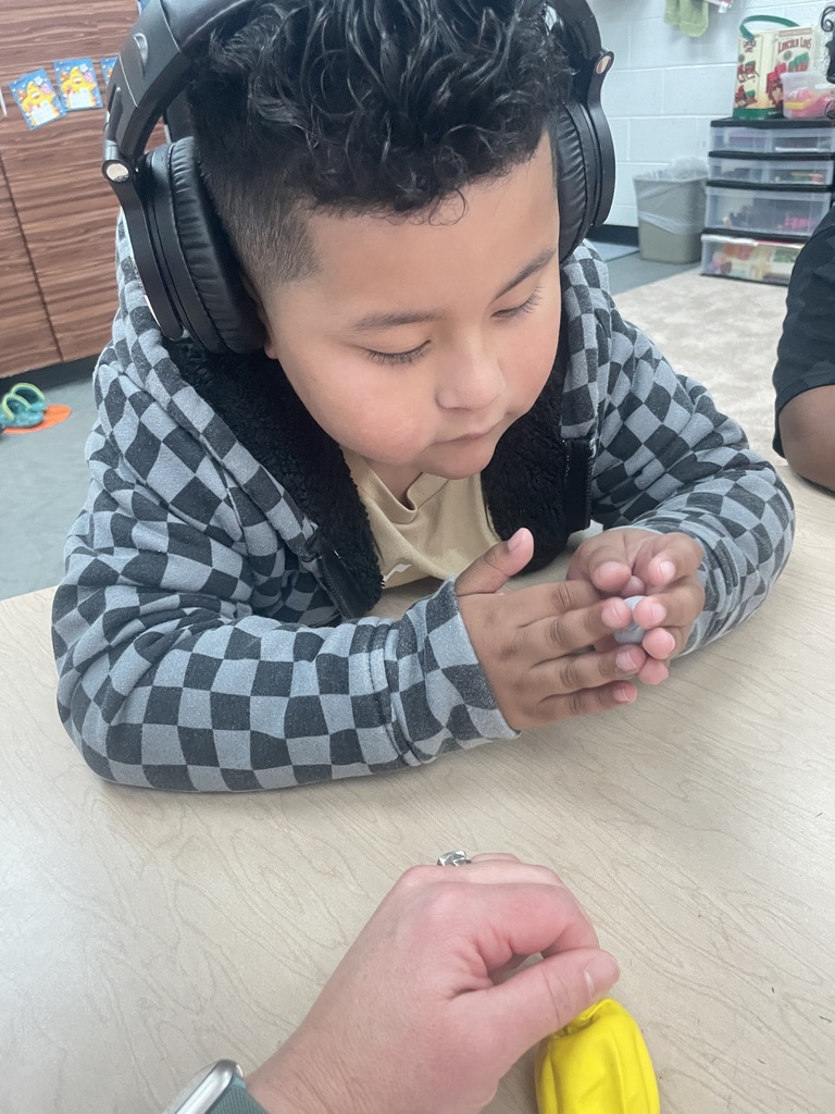 Child in headphones using a yellow object at a desk. A person holds the object.
