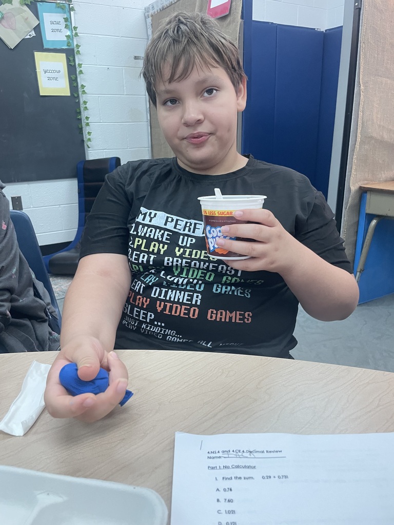 Child holding blue ball and cup at table, seated at desk with a paper in front.