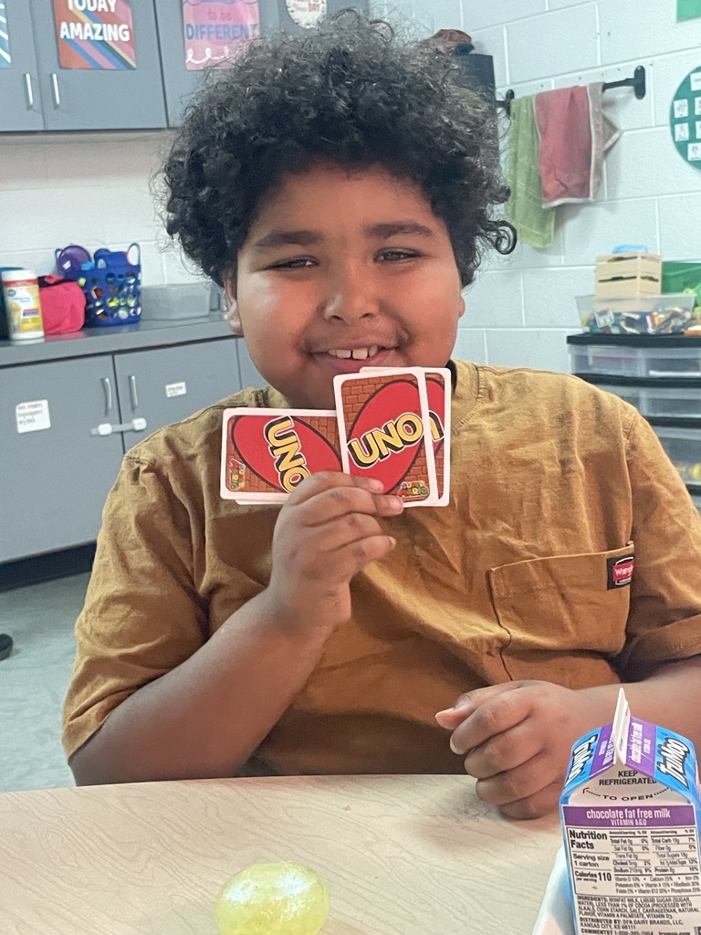 A student holds two cards, one labeled "UNO" and the other "ONE". A box of juice is on the table.