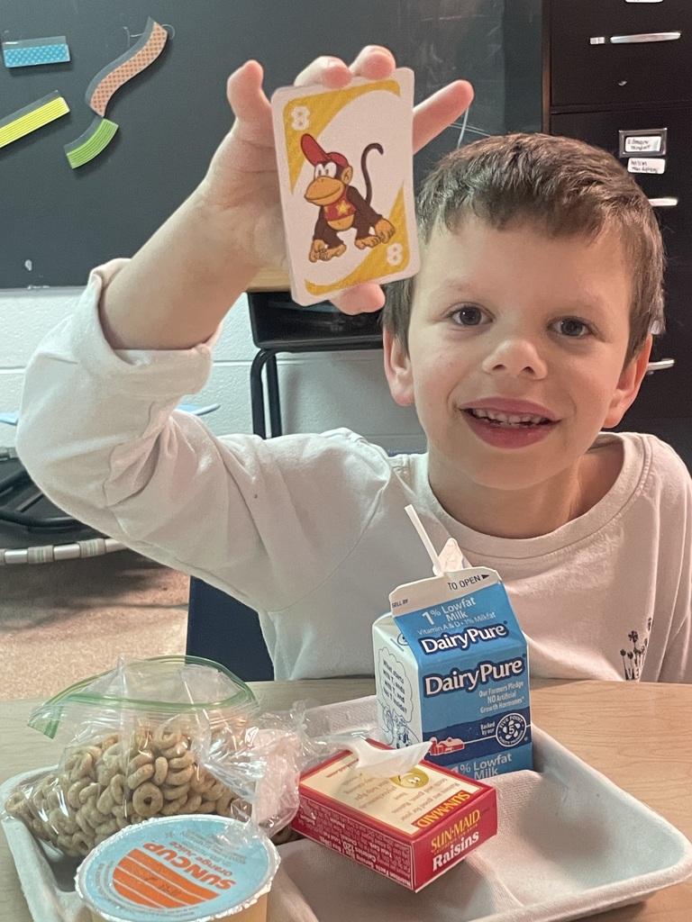 Child holding a card with a monkey, seated at a desk with a tray of food.