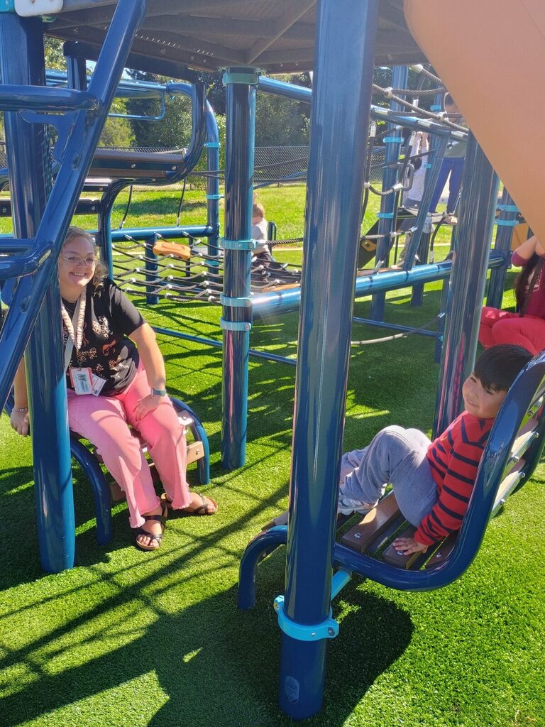 Two children sit on blue playground seats, one with pink pants, another in a striped shirt.