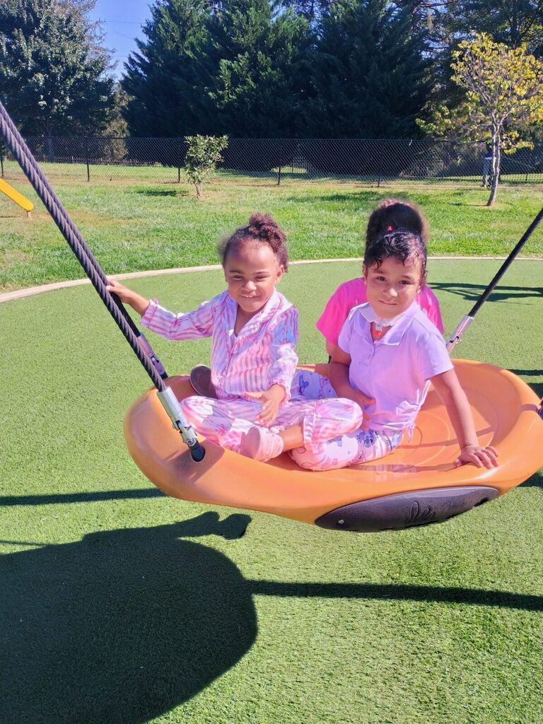 Two children sitting in a swing. One wears pink pajamas, and the other wears pink clothes. Trees and grass in background.