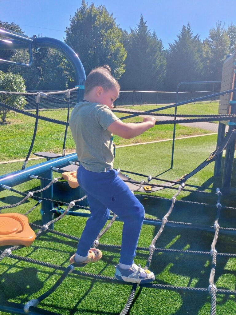 Child climbs blue net with ropes in outdoor playground on sunny day. Green grass, blue sky, and trees visible in background.
