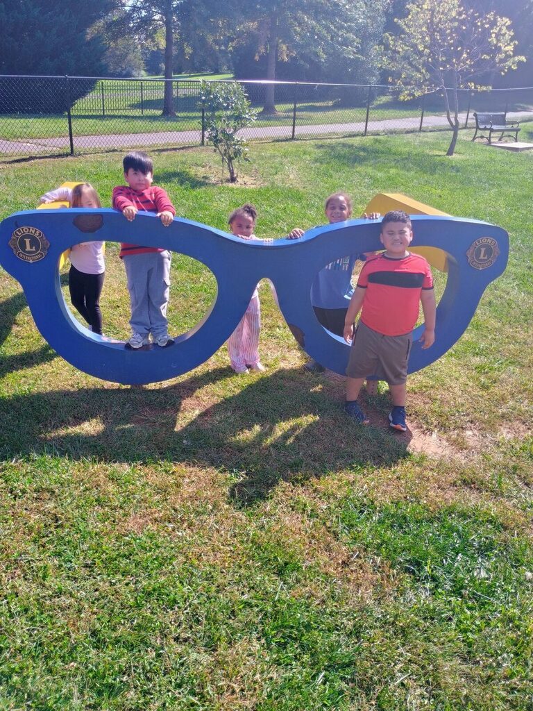 Six children stand behind a large blue object shaped like glasses in a grassy area.