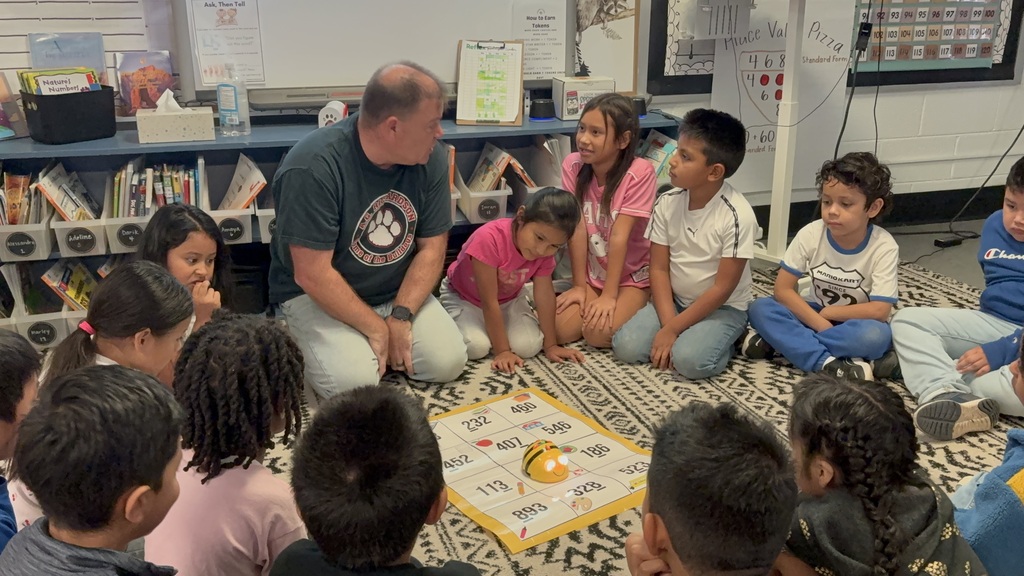 A group of children sit around a man in a classroom. He speaks to them from a kneeling position.