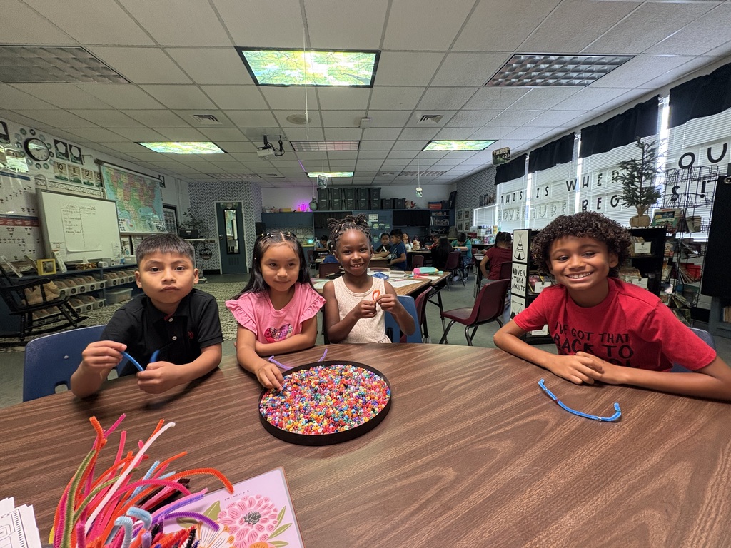 Four children seated around a table, smiling. Table contains beads and a colorful flower. Bright lighting, white walls, and ceiling with fluorescent lights.