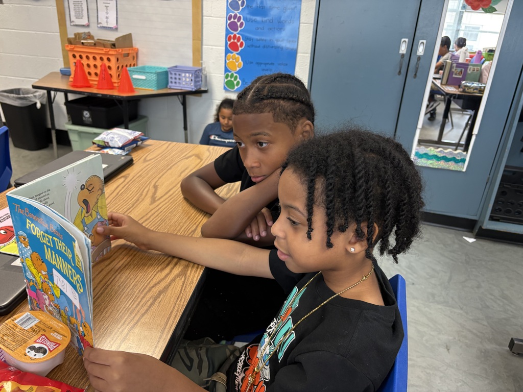 Two children at a table, one reading a book, the other looking at the book.