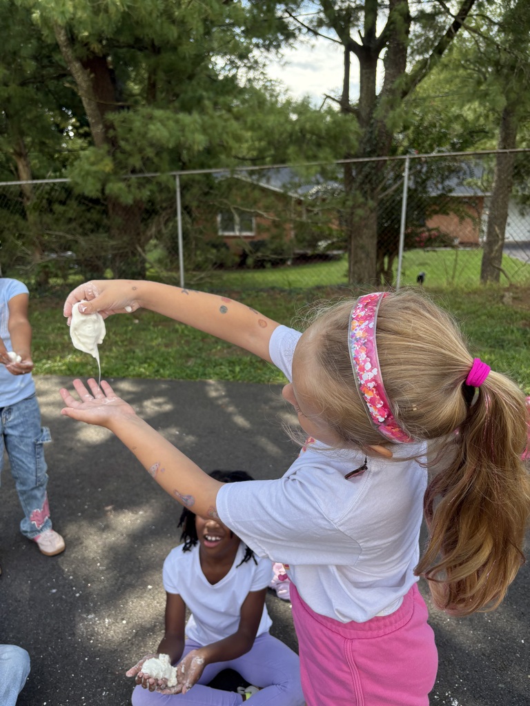 A girl holds a white substance and another girl sits nearby. Behind them, a fence, grass, and trees are visible.