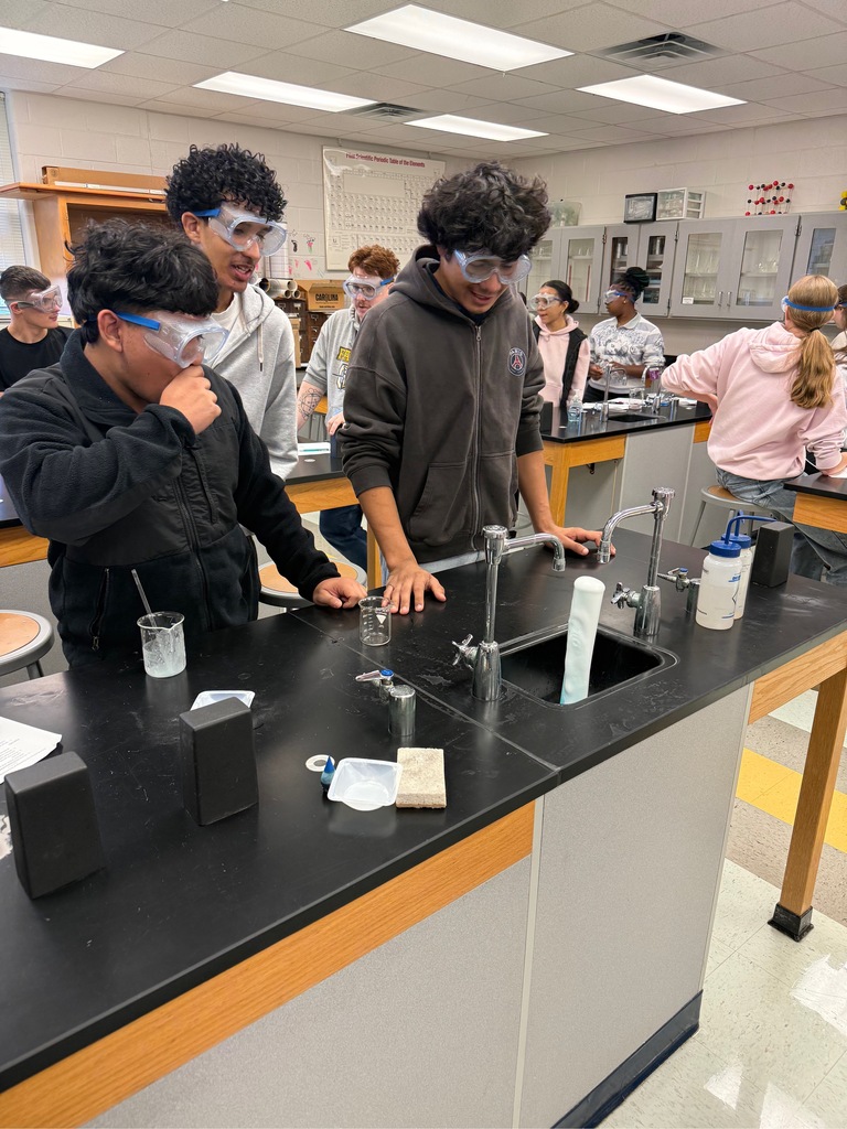 Students with safety goggles stand around a countertop in a classroom lab, working at sinks.