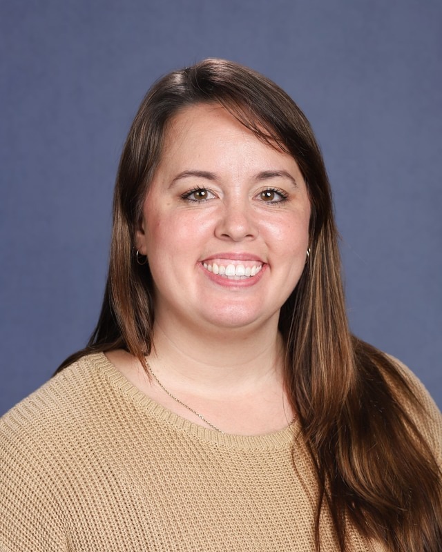 Woman with dark brown hair smiling at camera. Wearing cream sweater and a necklace. Blue background.