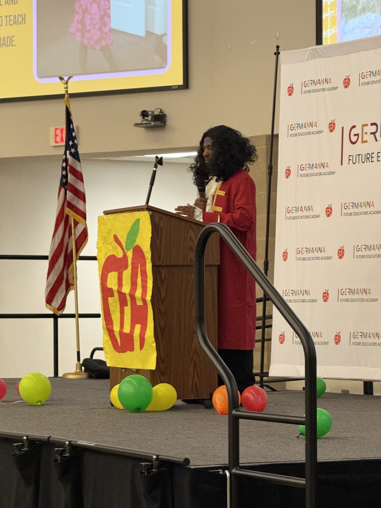 Person standing at a podium on stage with a US flag, yellow and green balloons.