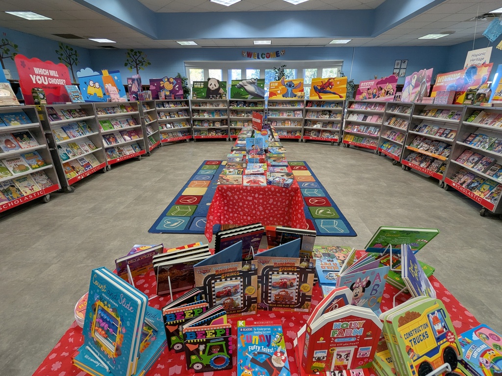 image shows the school library with the Scholastic book fair set up.  Cases of books in a U shape with tables up the middle.