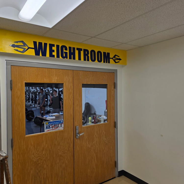 Entrance to weight room with a yellow sign above, wooden double doors with glass panels, and two people inside.