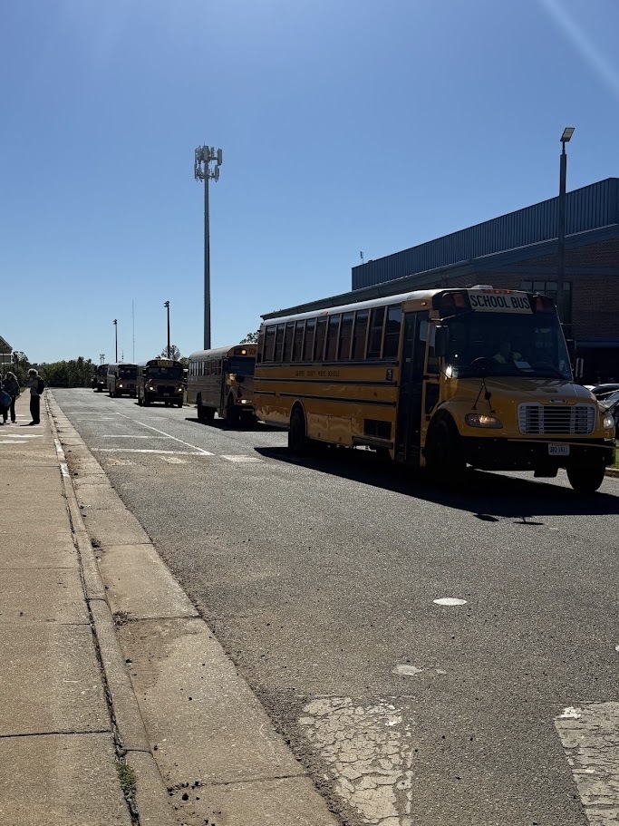 Several yellow school buses line up along the road outside Culpeper Middle School. A large bus fills the right side of the image, with additional buses extending into the distance. The school building, sidewalk, and bright blue sky provide the background.