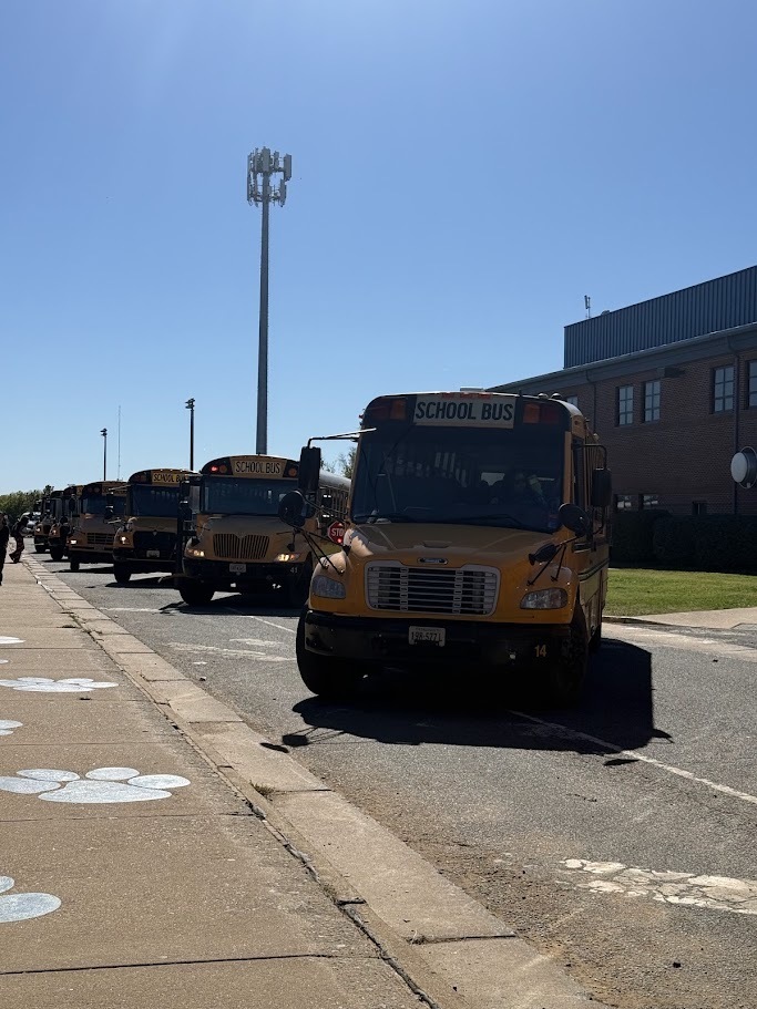 A row of yellow school buses lines the road outside Culpeper Middle School on a sunny day. The first bus faces the camera, with several additional buses lined up behind it. A brick school building, sidewalk, and clear blue sky are visible.