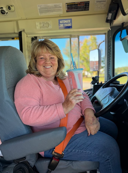 A smiling CMS school bus driver sits in the driver's seat of a yellow school bus, wearing a pink sweater and orange seat belt. She holds up a pink appreciation tumbler while posing for the camera, with the bus steering wheel and windshield visible beside her.