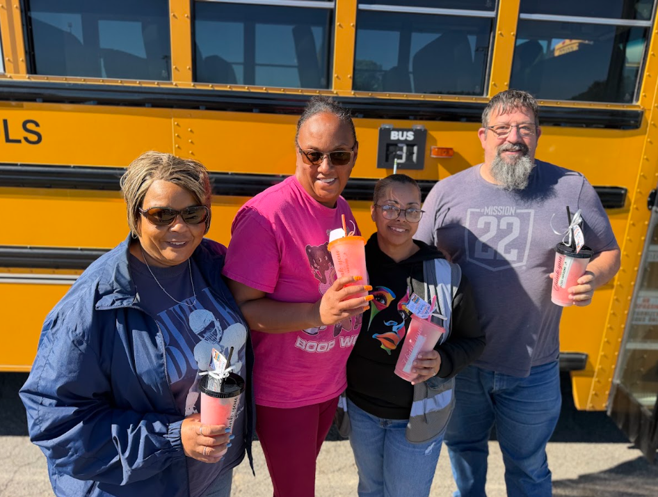 Four smiling CMS transportation professionals pose together in front of a yellow school bus on a sunny day. Three women and one man hold pink appreciation tumblers filled with treats. The group stands close together, celebrating Transportation Appreciation.