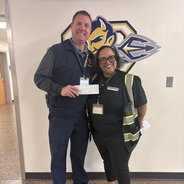 Two people stand in a hallway with a white wall, one holding a check and smiling.