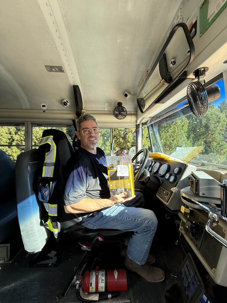 Man sitting in bus driver's seat holding book, safety vest hanging from back of chair.