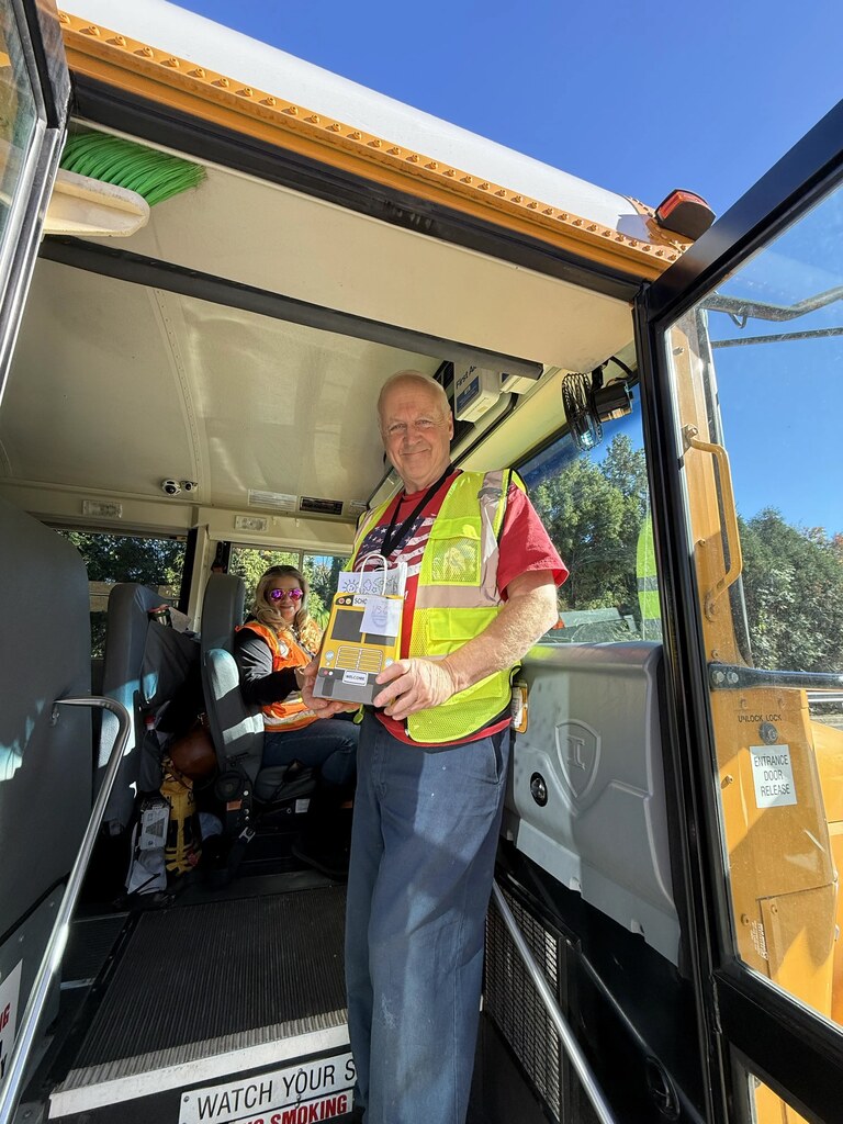 Adult man stands inside open door of school bus holding box while seated adult in yellow vest looks on.