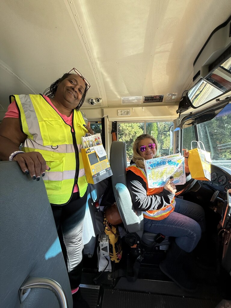 Two women in reflective vests in a bus. One holds a book and the other holds a bag.