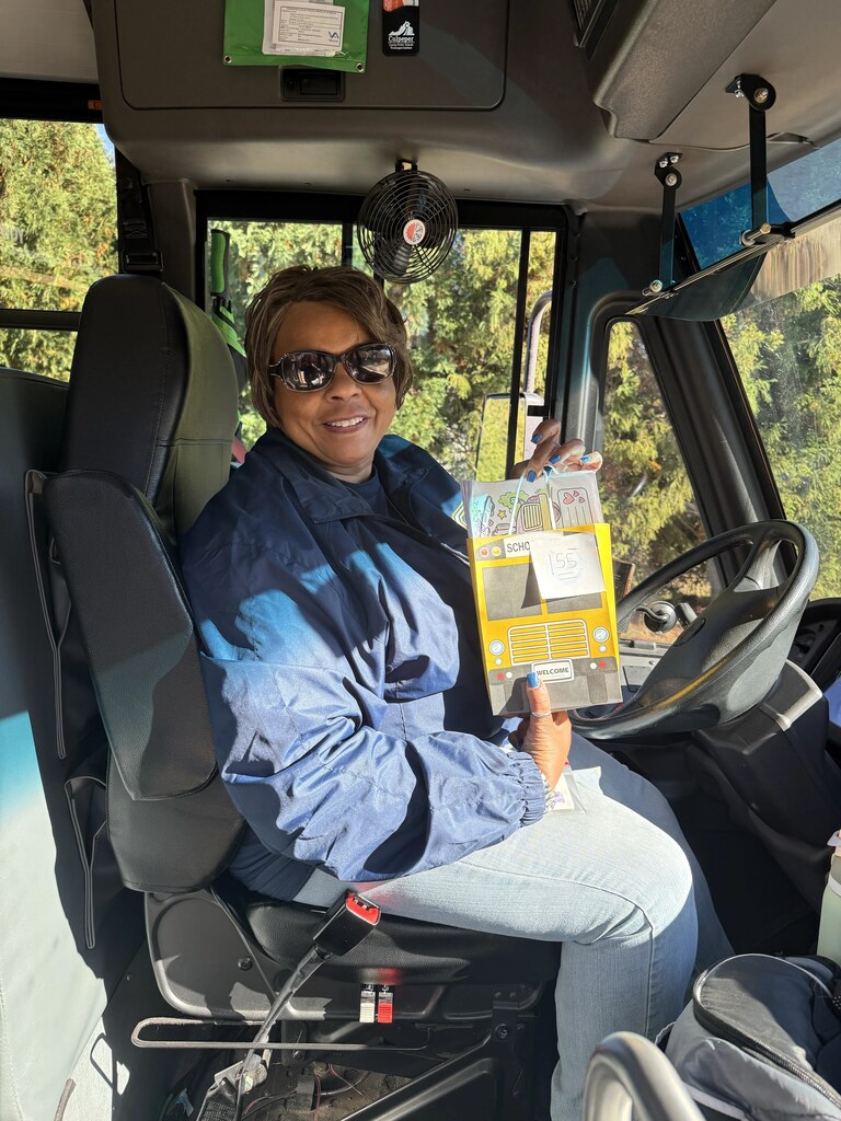 Bus driver seated, holding yellow book, wearing sunglasses, blue jacket, jeans, and a watch.