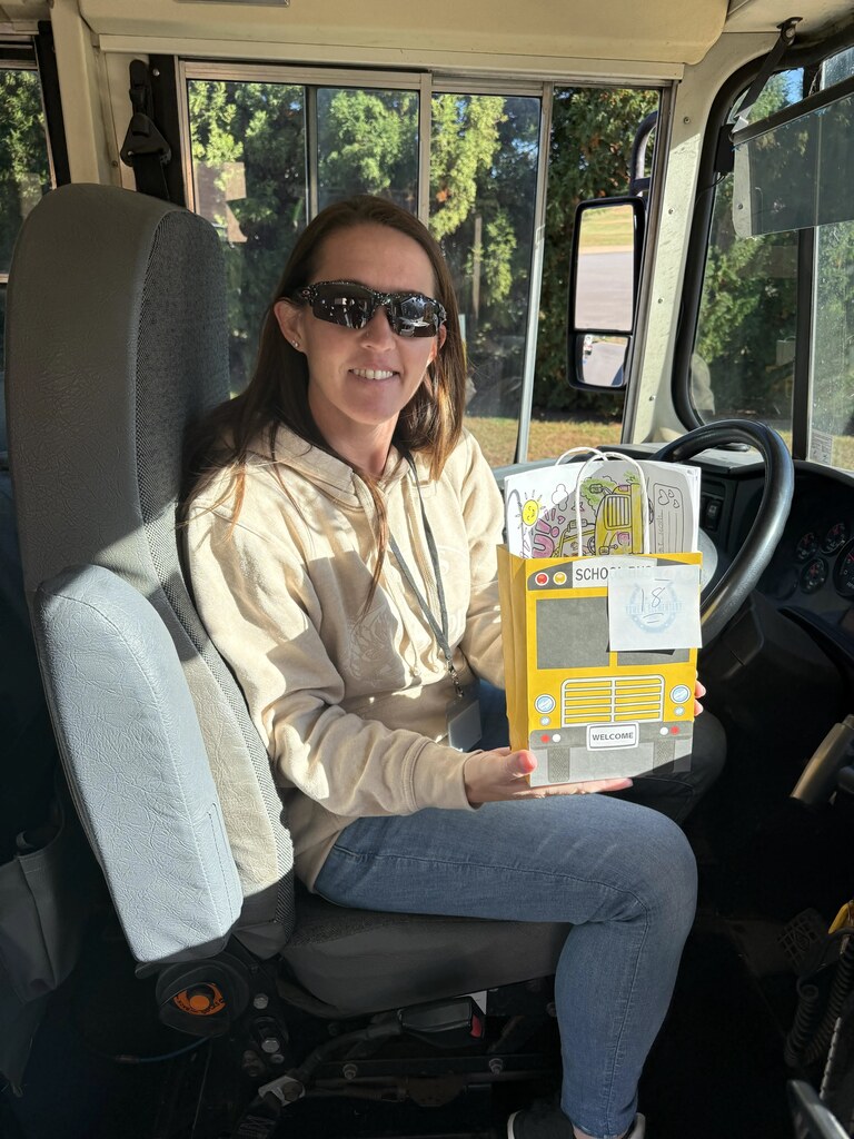 A woman sits in a bus driver's seat holding a yellow box. She wears sunglasses and a beige hoodie.