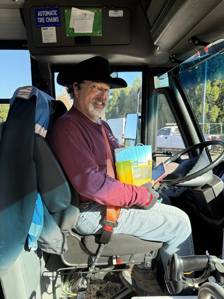 A man in a hat and glasses sits in a bus driver's seat, smiling, with a book.