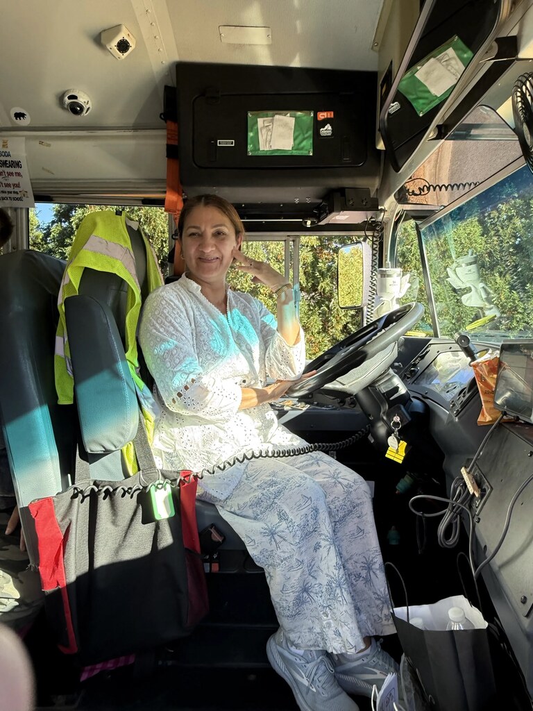 Woman seated in bus, wearing white pants, white shoes, and bracelet, with steering wheel and phone in hand.
