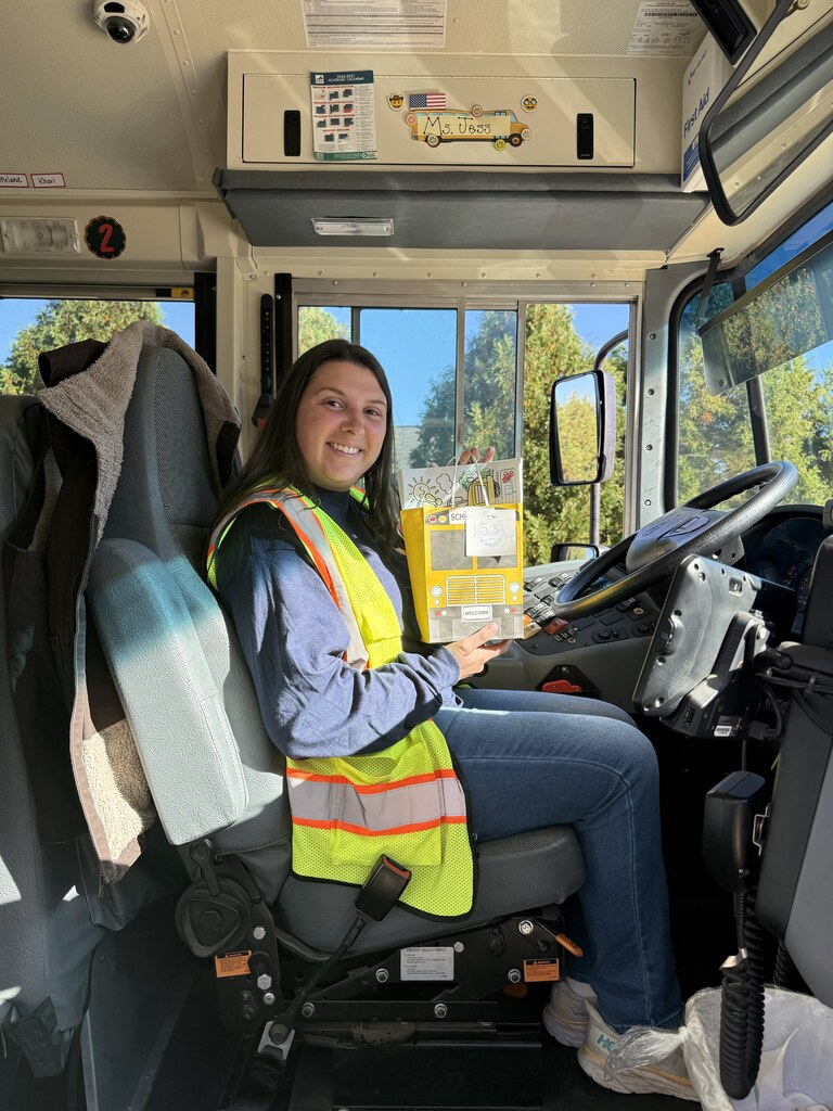 Woman wearing safety vest sits in driver's seat of bus with seat belt fastened.