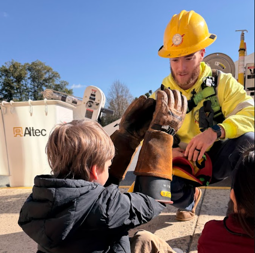 A man in a hard hat and safety vest helps a child put on a glove.