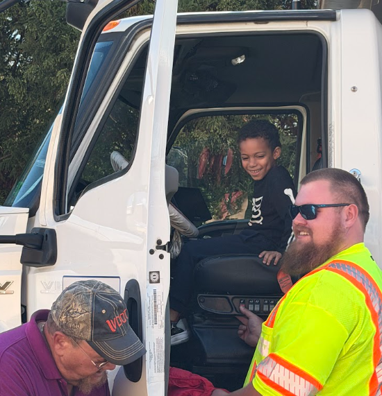 Man in reflective vest speaks with boy in vehicle; boy sits in driver's seat, smiling.