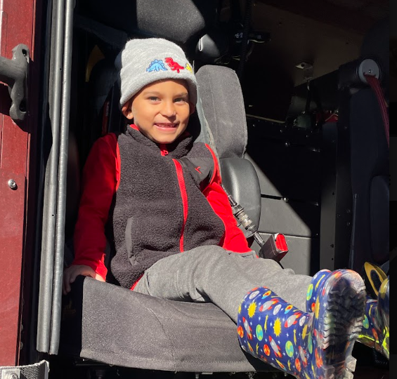 Child seated in fire truck chair, smiling, wearing red, gray, and blue clothing and a hat with a dinosaur.