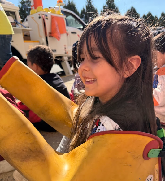 Child in yellow rain boot rides large yellow toy. Child smiles with others in background.