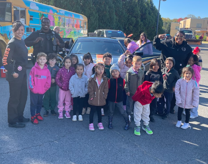 Police officers stand behind a car with children in front, some waving. Behind them are a school bus and a vehicle.