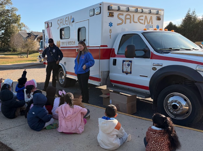 Students sit in a semicircle on a sidewalk, listening to an adult woman near an ambulance.