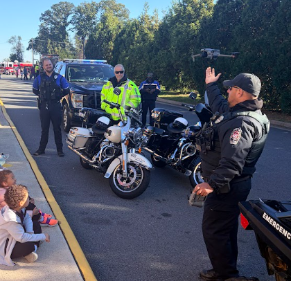 Police officers in uniform standing near children on a sidewalk, two motorcycles parked on the street, and a man waving.
