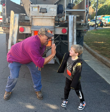 A man in a purple shirt bends to greet a boy in a jacket. A large truck is behind them.